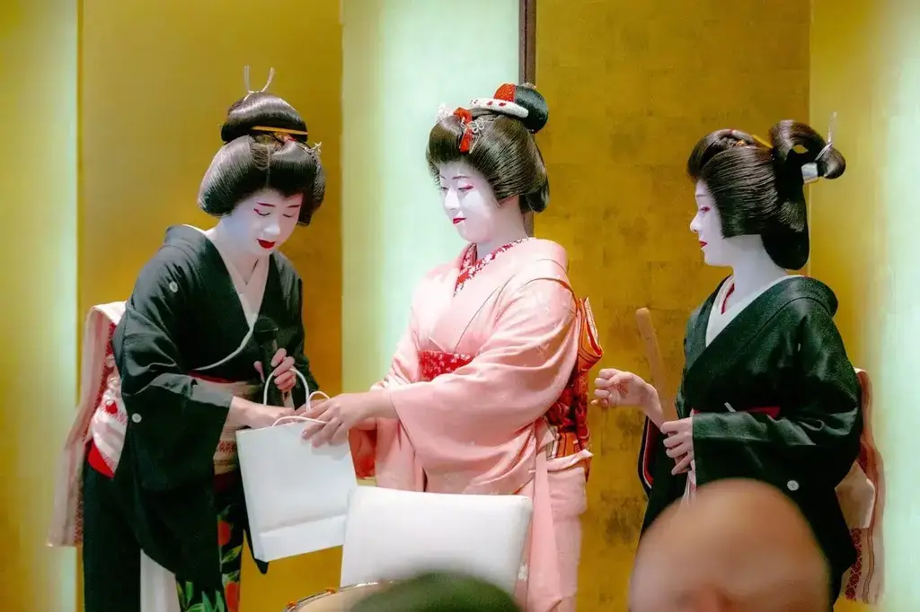 Three women wearing Japanese hairstyles and kimono. They also have white face makeup.