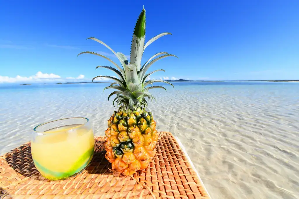 A pineapple on a beach next to pineapple juice.