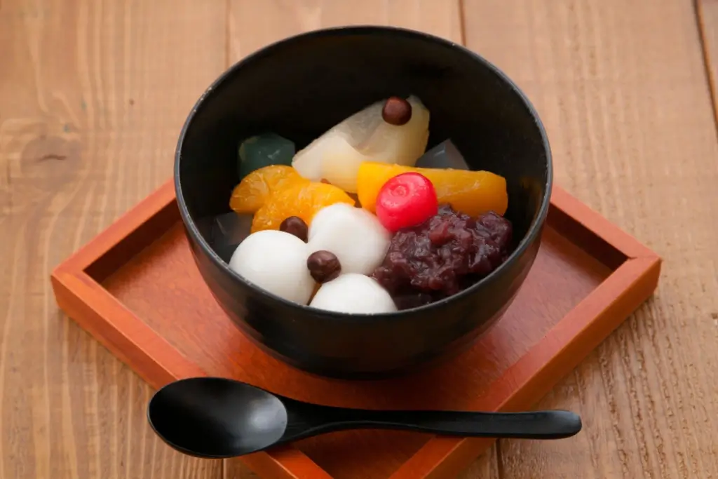 A bowl of anmitsu with shiratama, fruit, red bean paste and agar jelly inside.